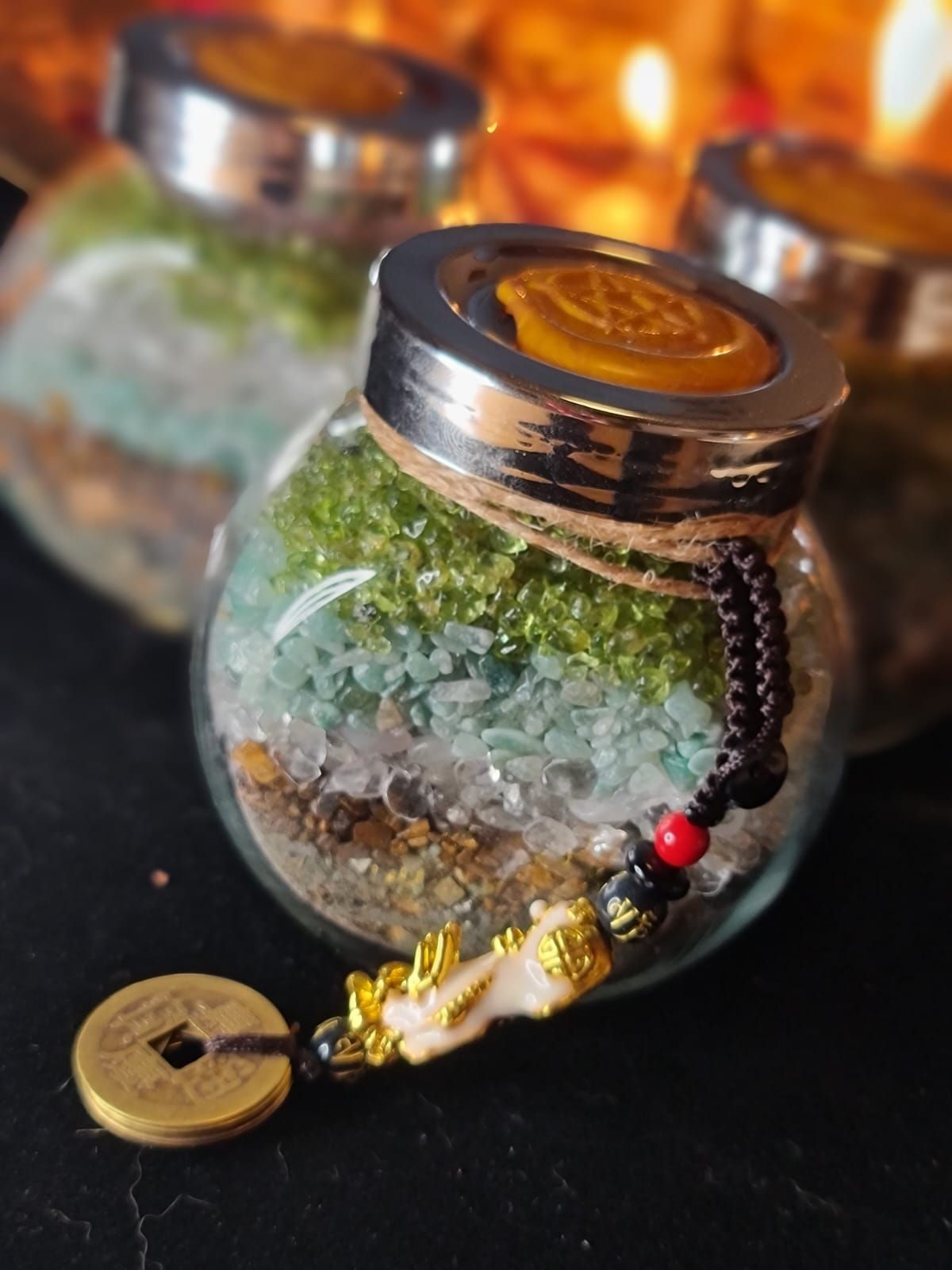 Glass jar filled with layered crystals and topped with a coin and decorative beads on a black surface.